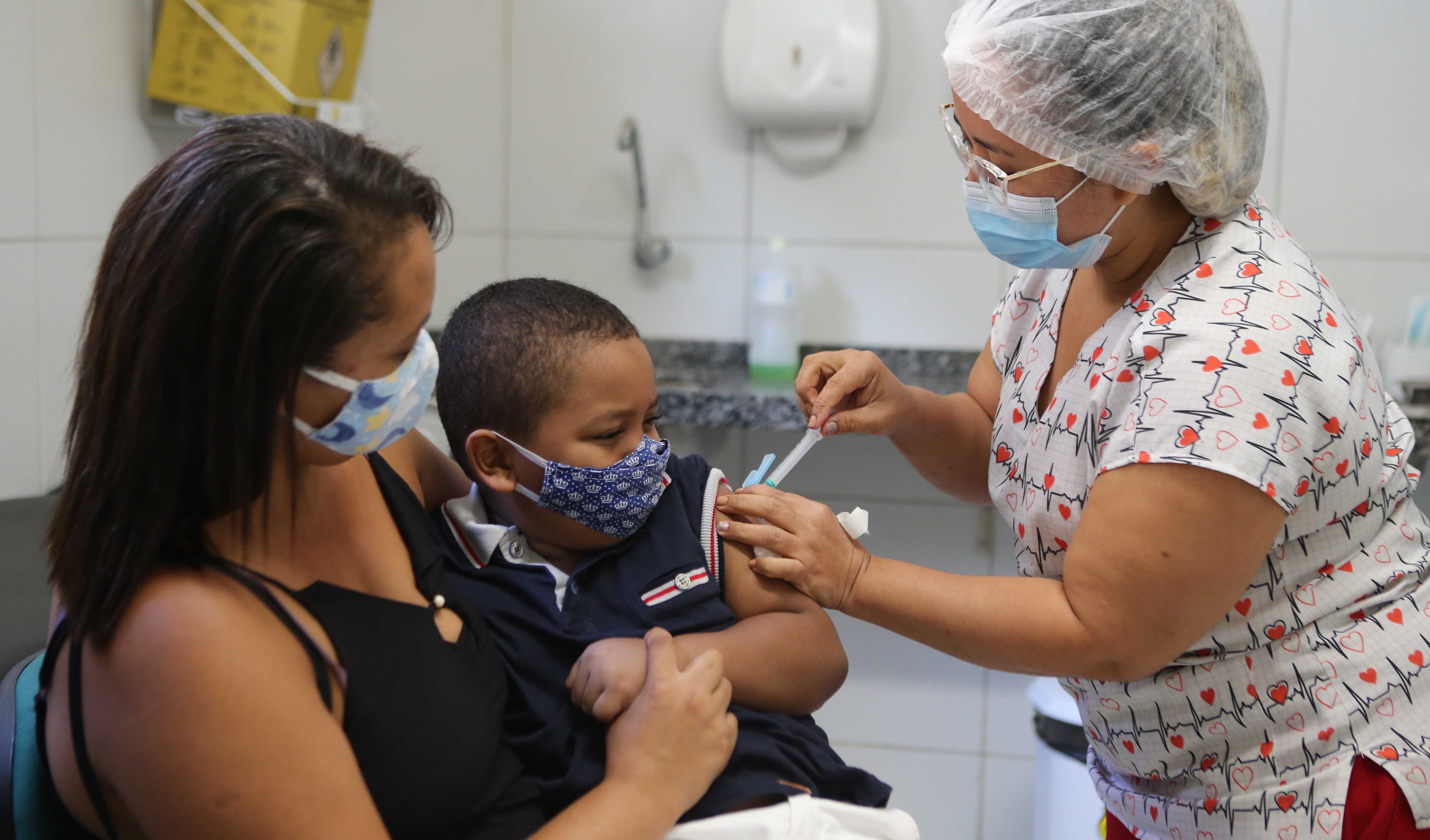 uma enfermeira vacina uma criança que está no colo da mãe. Todas usam máscaras
